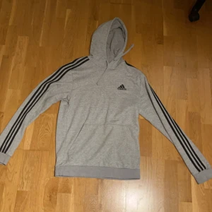 Grå hoodie från Adidas - Säljer en grå hoodie från Adidas med de klassiska tre ränderna längs ärmarna. Tröjan har en justerbar huva och en stor ficka framtill. Perfekt för en avslappnad stil. 🖤.                                                Tar emot byten