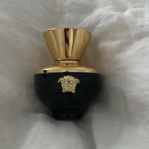 Svart/marinblå och guld parfymflaska från Versace - Elegant parfymflaska i svart/marinblå och guld från Versace. Flaskan har en lyxig design med det ikoniska Medusa-emblemet i guld på framsidan. Den har en rund form med en bred, guldig kork som ger en exklusiv känsla. En lyxig och mogen doft, 50 ml, knappt använd