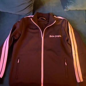 Svart hoodie från Palm Angels - Säljer en svart hoodie från Palm Angels med vita ränder längs ärmarna. Den har dragkedja framtill och Palm Angels-logga på bröstet. Den är knappast använd på grund av att den var förstor. Inga hål eller fläckar. Alla qr coden funkar såklart.