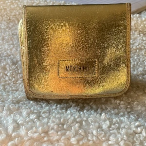 Guldig plånbok från Moschino - Säljer en snygg guldig plånbok från Moschino Den har en glansig yta och en praktisk design som passar perfekt i väskan. Perfekt för att hålla ordning på kort och kontanter. En stilren accessoar för den som gillar att sticka ut.