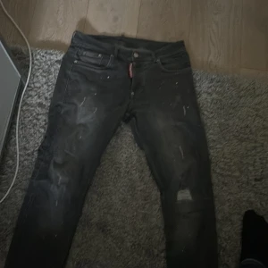 Dsquared2 jeans - Säljer ett par svarta jeansbyxor med slitningar och en klassisk femficksdesign. Byxorna har en knappgylf och en röd etikett vid linningen. Perfekta för en avslappnad stil.