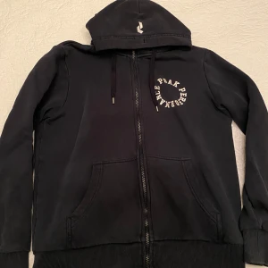 Mörkblå hoodie från Peak Performance - Säljer en mörkblå hoodie från Peak Performance med dragkedja och broderad logga på bröstet. Den har en praktisk huva och fickor framtill. 