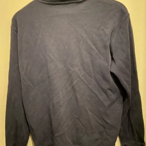 Mörkblå tröja med dragkedja från Ralph Lauren - Säljer en mörkblå tröja från Ralph Lauren med dragkedja och röd broderad logotyp på bröstet. Tröjan har långa ärmar och en klassisk krage. Perfekt för en stilren look.