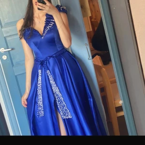 Blå långklänning med paljettdetaljer - Elegant blå långklänning i satin med djup v-ringning och knytband i midjan. Klänningen har en slits framtill och är dekorerad med glittrande paljetter längs halsringningen och knytbandet.
