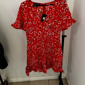 Röd blommig klänning från Parisian - Charmig röd klänning från Parisian med blommigt mönster. Den har korta ärmar med volangdetaljer och en söt volangkant längst ner. Perfekt för en somrig look!