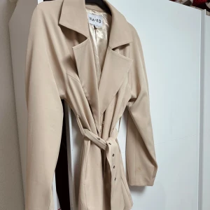 Beige trenchcoat från NA-KD - Snygg beige trenchcoat från NA-KD med knytbälte i midjan och klassisk krage. Perfekt för övergångsperioder med sin stilrena design och långa ärmar. Passar perfekt för s/m/l storlek