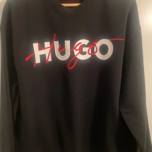 Svart sweatshirt från Hugo - Snygg svart sweatshirt från Hugo med stort tryck i vitt och rött på framsidan. Tröjan har långa ärmar och en klassisk rund halsringning. Perfekt för en stilren och avslappnad look.