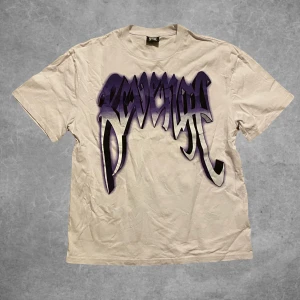Vit t-shirt med lila tryck från Revenge - Cool vit t-shirt från Revenge med ett stort lila tryck fram och bak. Perfekt för dig som vill sticka ut med en unik design. T-shirten har korta ärmar och en normal passform.