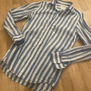 Linneskjorta från Massimo Dutti - Snygg blå- och vitrandig skjorta från Massimo Dutti i 100% linne. Perfekt för en stilren look med knappar framtill och klassisk krage. Skjortan har långa ärmar och en avslappnad passform.                -Köpt på Massimo Dutti butik i Stockholm               -Superbra passform, för både herr och dam 