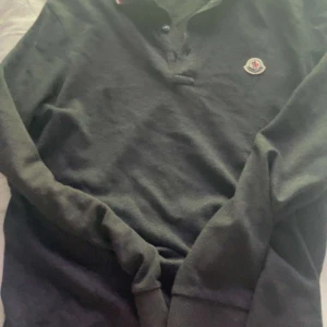 Svart pikétröja från Moncler - Snygg svart pikétröja från Moncler med klassisk krage och knappar framtill. Tröjan har Monclers logotyp broderad på bröstet och en stilren design med ribbade muddar vid ärmsluten.