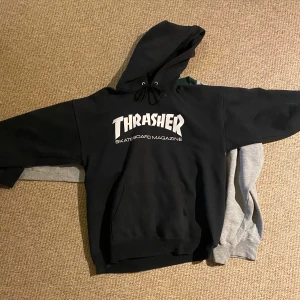 Svart hoodie från Thrasher - Säljer en svart hoodie från Thrasher med det ikoniska logotyptrycket på framsidan. Tröjan har en stor ficka framtill och en justerbar huva med snören. Perfekt för en avslappnad stil.