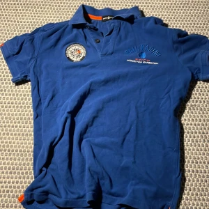 Blå pikétröja från Sail Racing - Säljer en blå pikétröja från Sail Racing med broderad logga och text 'Antarctica Expedition' på bröstet. Tröjan har korta ärmar och en klassisk krage med knappar. Perfekt för en avslappnad stil.