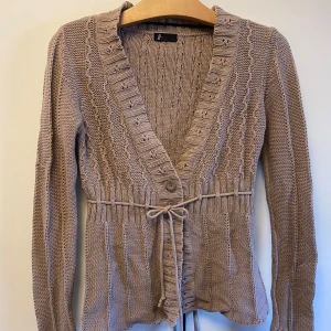 Vero Moda Brun Beige stickad kofta strl M - Säljer en snygg beige stickad kofta från Vero Moda i storlek M. Koftan har en v-ringad design med knytband i midjan och ribbade detaljer längs framstycket. Koftan är mycket väl omhändertagen. 