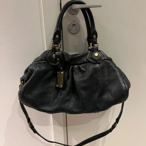 Marc Jacobs väska  - Snygg svart handväska i skinn med justerbar axelrem och guldfärgade detaljer. Väskan har en rymlig insida med ett mönstrat foder och dragkedja. Perfekt för att bära med sig alla nödvändigheter med stil.