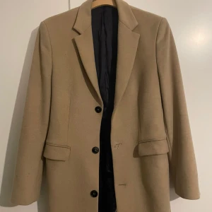 Beige kappa från Zara  - Säljer en stilren beige kappa från Zara Man. Kappan har en klassisk design med knappar framtill och två fickor. Perfekt för höst och vår.