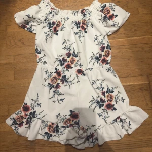 Vit blommig byxdress med volangdetaljer - Charmig vit byxdress med blommönster i rosa och grönt. Byxdressen har korta ärmar och volangdetaljer vid fållen. Perfekt för sommarens varma dagar.