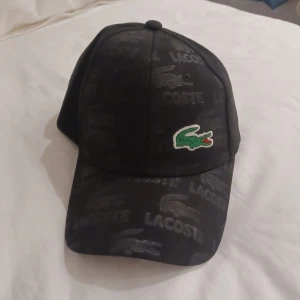 Svart keps från Lacoste - Snygg svart keps från Lacoste med broderad grön krokodil på framsidan. Kepsen har justerbart spänne i metall baktill och märkeslogga både inuti och utanpå. Perfekt för en stilren look. Perfekt för en golfare eller något liknande. Fick den i present vet ej ifall den är äkta