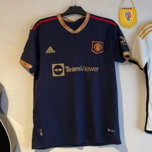 Blå Manchester United fotbollströja - Snygg blå Manchester United tröja från Adidas med guldiga detaljer och klubbens emblem på bröstet. Tröjan har korta ärmar och Premier League-märke på ärmen. Perfekt för fans som vill visa sitt stöd.