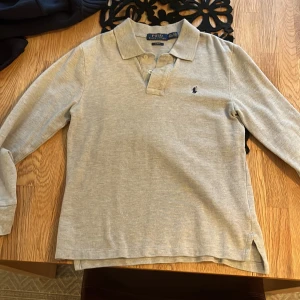 Grå pikétröja från Ralph Lauren - Säljer en stilren grå pikétröja från Ralph Lauren med klassisk krage och knappar. Tröjan har långa ärmar och en liten broderad logotyp på bröstet. Perfekt för en avslappnad men ändå snygg look.Storlek 14-16 år