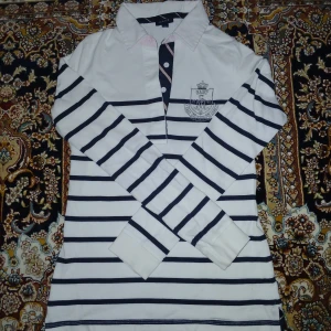 Vit och svart randig pikétröja - Snygg vit och svart randig pikétröja med långa ärmar och knappar framtill. Tröjan har en broderad detalj på bröstet som ger en stilren look. Perfekt för en avslappnad men ändå uppklädd stil.