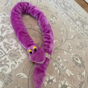 Mjukis orm  - Mysig och lekfull ormformad  orm  i lila plyschmaterial. .ormen  har stora gula ögon och en röd tunga som sticker ut.den sätteri fast på sin svans ,som barnen kan hänga rund  axlar eller hur dem vill . Svensk tillverkat frå år 1988 . Maskintvätt 30grader så bra kvalitet . Perfekt som en rolig inredningsdetalj eller som en mjuk leksak.längd är 1,24cm