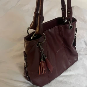 Trendig mörkröd väska i nyskick - Rymlig väska i faux läder, aldrig använd då den inte kommit till användning. Levererades med något unken lukt, men går att få bort med rengöringsmedel. Rymmer dator (21x30cm), mappar mm💕 Inköpspris: 450 kr 