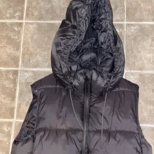 Svart dunväst från Zara - Snygg svart dunväst från Zara med dragkedja och huva. Perfekt för kyligare väder och ger en stilren look. Västen är quiltad och har en normal passform.