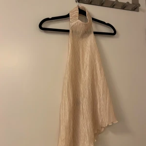 Beige asymmetrisk topp från Zara - Snygg beige topp från Zara med asymmetrisk design och hög hals. Toppen har en elegant plisserad struktur och är ärmlös, vilket ger en stilren look. Perfekt för en chic och modern stil.