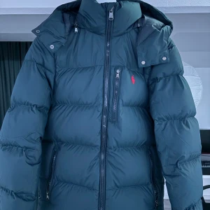 Mörkblå dunjacka från Polo Ralph Lauren - Säljer en snygg mörkblå dunjacka från Polo Ralph Lauren. Jackan har en dragkedja framtill och praktiska fickor med dragkedjor. Den är långärmad och perfekt för kyliga dagar. Den ikoniska loggan syns på bröstet. Perfekt för dig som vill hålla stilen i vinter! ❄️