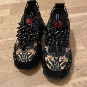 Versace chain reation 44  - Snygga svarta sneakers med unikt mönster i guld och orange. Skorna har en tjock sula och snörning med svarta och vita skosnören. Perfekta för en trendig look.