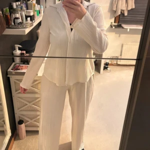 Vit plisserad skjorta och byxor - Säljer en stilren vit plisserad skjorta med matchande byxor. Skjortan har långa ärmar och knappar framtill, perfekt för en elegant look. Byxorna är vida och bekväma, vilket ger en avslappnad men chic stil.