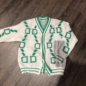 Vit och grön kofta med geometriskt mönster - Säljer en unik vit kofta med grönt geometriskt mönster. Koftan har långa ärmar och knappar framtill. Perfekt för att sticka ut med en retro look!