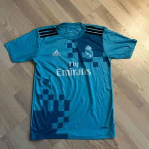Blå Real Madrid fotbollströja från Adidas - Säljer en blå Real Madrid fotbollströja från Adidas med korta ärmar och Climacool-material. Tröjan har ett pixelmönster i mörkare blått och texten 'Fly Emirates' på framsidan. På baksidan finns namnet 'Ronaldo' och nummer 7 tryckt i vitt.