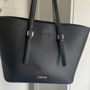   Calvin Klein handväska  - Fin CK handväska i svart läder som är använd ett fåtal gånger. Inga skador eller slitage.  Magnetknäppe samt löstagbar innerväska med dragkedja. Mått: H25cm, B42cm, D16cm