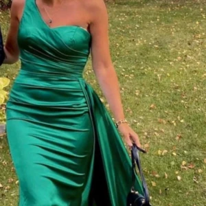 Grön långklänning med slits - Säljer en fantastisk grön långklänning i satin med en elegant slits. Klänningen har en oneshoulder-design som ger en stilren look. Perfekt för fest eller bal!