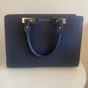 Michael kors - Marin blå favvo väska vill ha nytt hem. Fina gulddetaljer och i fint skick. Väldigt rymlig, ända som syns på användning är fötterna (se bild) knappt något att se.