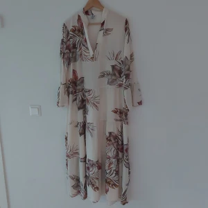 Blommig långklänning från Stajl - Säljer en superfin långklänning från Stajl med ett härligt blommigt mönster. Klänningen är i en ljus färg med långa ärmar och en v-ringad design. Perfekt för både sommar och vår, och den har en skön och luftig passform.