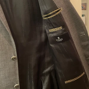 Grå kavaj från Tiger of Sweden - Säljer en stilren grå kavaj från Tiger of Sweden. Kavajen har en klassisk passform med två knappar fram och fickor med lock. Den är fodrad med ett elegant svart tyg och har fina detaljer som knappar vid ärmsluten. Perfekt för både formella och semi-formella tillfällen.