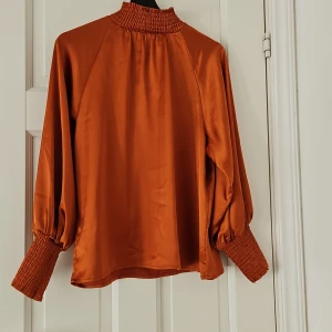 Orange satinblus med smockdetaljer - Säljer en superfin orange/ blus i satinmaterial. Den har långa ärmar med smockdetaljer vid ärmslut och hals, vilket ger en snygg och unik look. Perfekt för både till vardags och fest. Oanvänd från By Malina