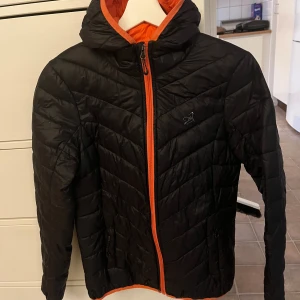 Svart och orange pufferjacka från 2117 of Sweden - Säljer en snygg svart pufferjacka med orange foder från 2117 of Sweden. Jackan har en dragkedja framtill och en mysig huva. Perfekt för kyliga dagar och ger en cool kontrast med det orangea fodret. Den är quiltad och har en normal passform. Perfekt för höst och vinter!