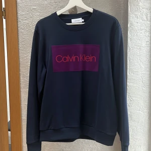 Mörkblå sweatshirt från Calvin Klein - Säljer en stilren mörkblå sweatshirt från Calvin Klein i storlek L. Tröjan har en stor lila logga på bröstet och är i mycket bra skick. Perfekt för både vardag och avslappnade tillfällen. Skön och enkel att matcha med det mesta!