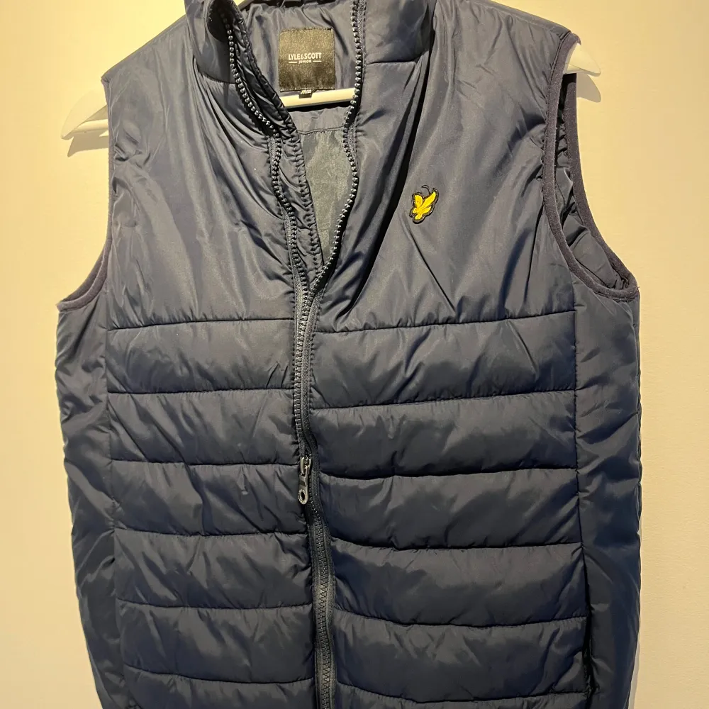 Säljer en snygg blå dunväst från Lyle & Scott. Västen har en dragkedja framtill och en broderad logga på bröstet. Perfekt för lager-på-lager under höst och vår. Den är i mycket bra skick och passar både till vardags och lite finare tillfällen.. Jackor.