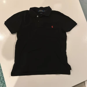 Svart pikétröja från Ralph Lauren - Säljer en snygg svart pikétröja från Ralph Lauren i slim fit. Den har en klassisk krage och korta ärmar, med det ikoniska röda logotypbroderiet på bröstet. Perfekt för både vardag och fest! 🖤