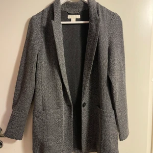 Grå kavaj från H&M - Snygg grå kavaj från H&M i ett klassiskt fiskbensmönster. Perfekt för både kontoret och mer avslappnade tillfällen. Den har långa ärmar och en enkel knapp framtill. 