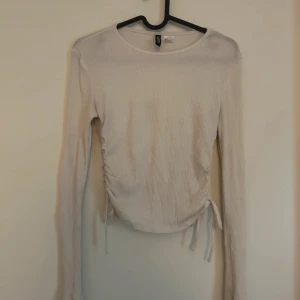 ribbad tröja med knyt - Säljer en superfin vit ribbad topp med långa ärmar. Den har en snygg knytning på sidan som ger en cool detalj och gör den lite croppad. Perfekt för både vardag och fest!