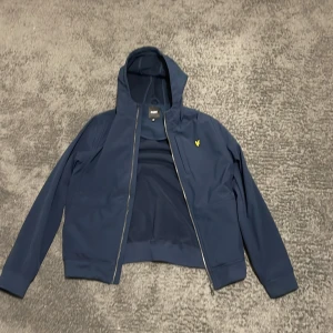 Blå jacka med huva från Lyle & Scott - Säljer en snygg blå jacka från Lyle & Scott med huva och dragkedja. Perfekt för vår och höst! Den har en gul logga på bröstet och är i mycket bra skick. Passar både till vardags och lite finare tillfällen. Den har två innerfickor och en på bröstet.