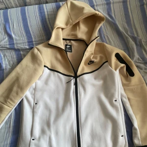 Beige och vit Teach från Nike - Säljer en snygg beige och vit teach från Nike i storlek M. Den har en dragkedja framtill och en praktisk huva. Perfekt för höst och vår, med en stilren design och bekvämt material.