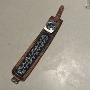 Säljer ett coolt vintage armbandsur med ett unikt mönstrat läderarmband. Från Sapmi. Klockan har en silverfärgad boett och en mörk urtavla. Armbandet är brunt med ett blått och silverfärgat mönster. Perfekt för dig som gillar retrostil!