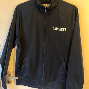 Säljer en marin blå track jacket från Carhartt i storlek M. Jackan är i 100% polyester och har en dragkedja framtill. Perfekt för höst och vår med sin stilrena design och bekväma passform. Passar både till vardags och lättare träning.