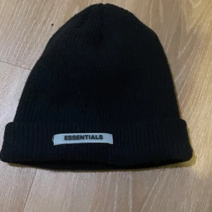 Svart stickad mössa från Essentials - Säljer en stilren svart stickad mössa från Essentials. Den är perfekt för kalla dagar och har en enkel design med en uppvikt kant och en diskret logga framtill. Passar till alla outfits och håller dig varm hela vintern.